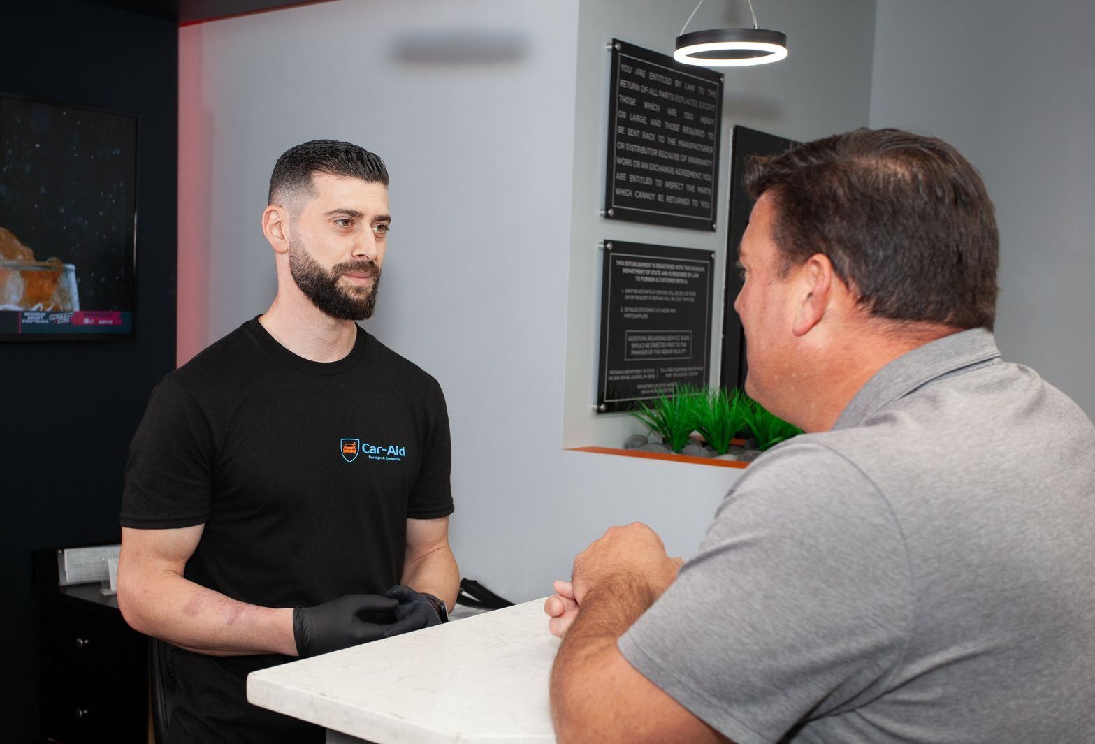 A man in a black shirt with a logo talks to a customer in a gray shirt at a counter in a modern office.