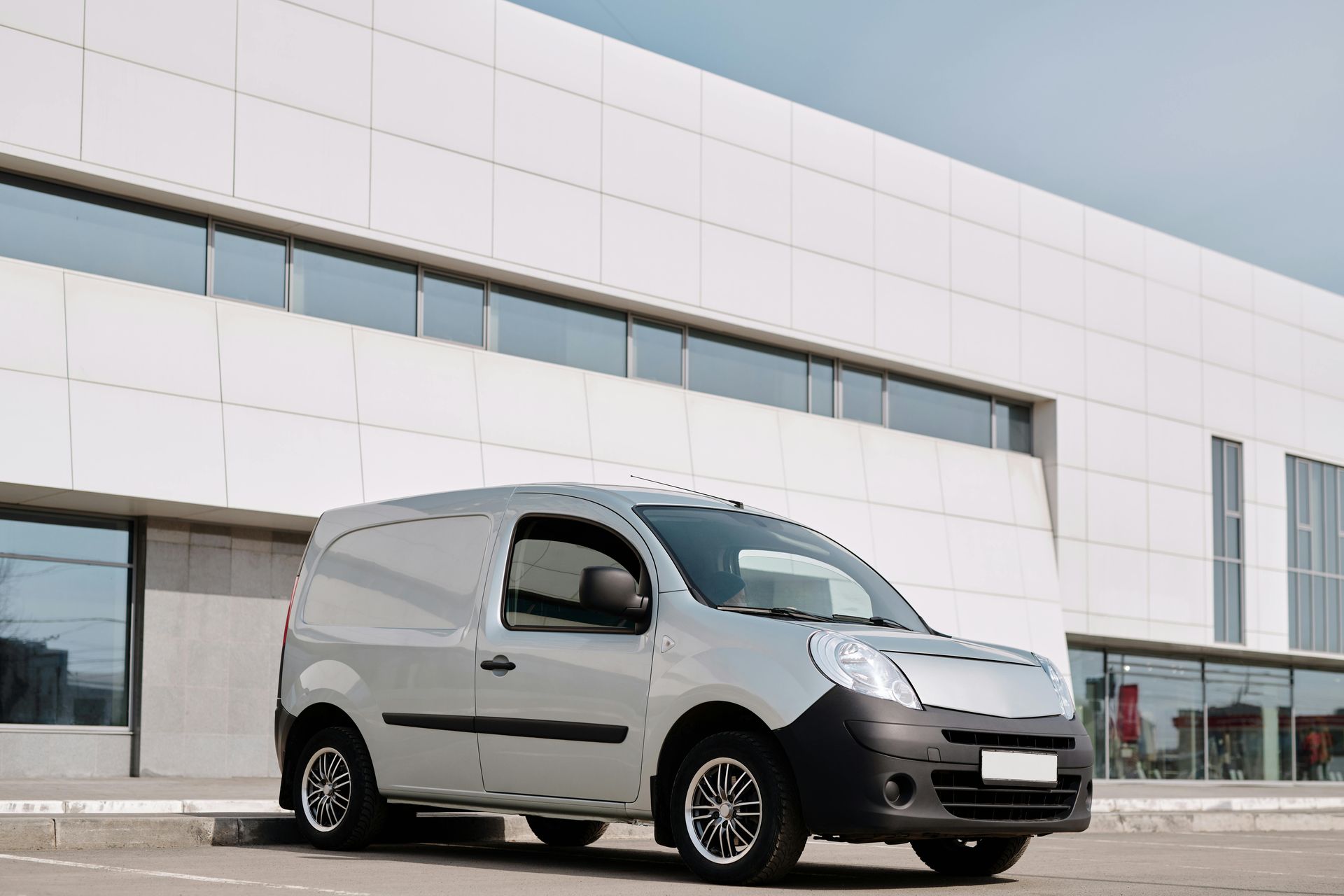 Silver cargo van parked in front of a white building with large windows.