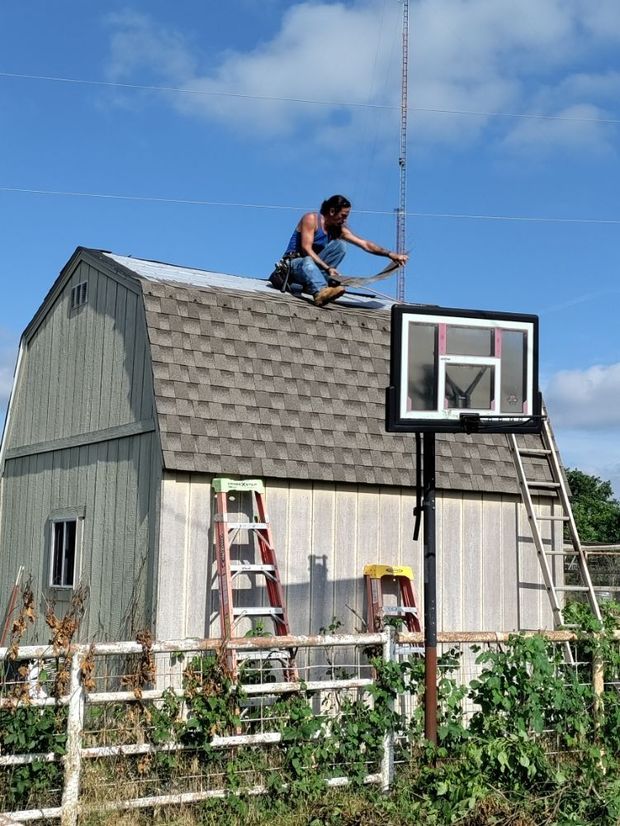 A man is working on the roof of a barn next to a basketball hoop.