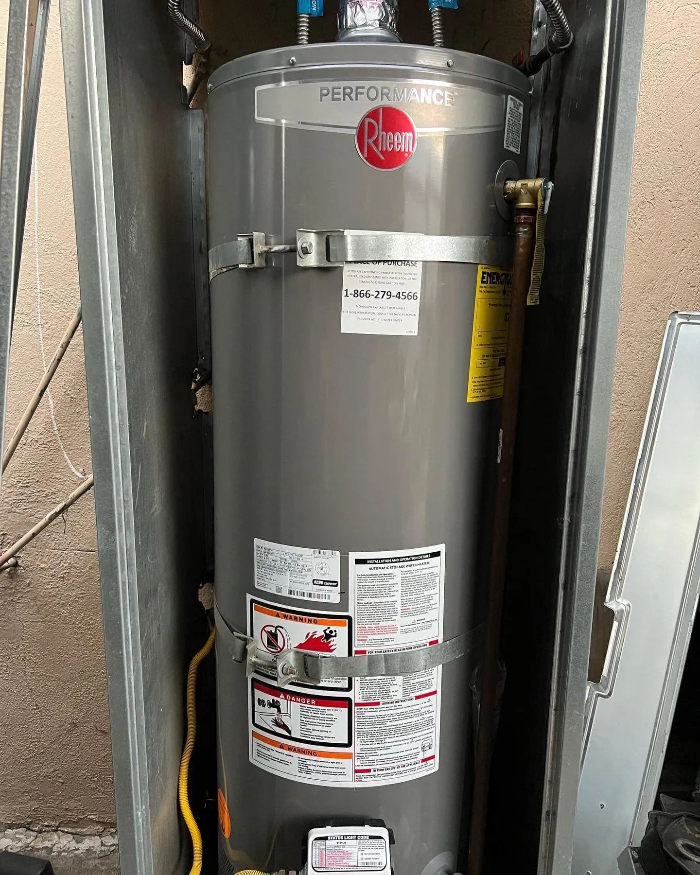 Gray Rheem water heater, encased in metal, with various labels and fittings, against a brick wall.