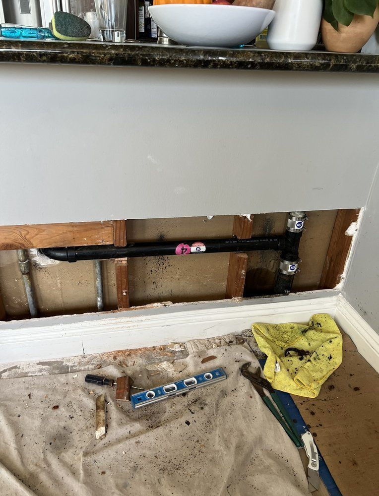 Plumbing repair: Black pipe exposed in a wall. Pink tape marks the pipe. Tools and debris on the floor below.