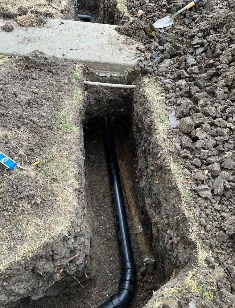 A black plastic pipe sits inside a freshly dug trench next to a concrete sidewalk, with dirt piles on either side.