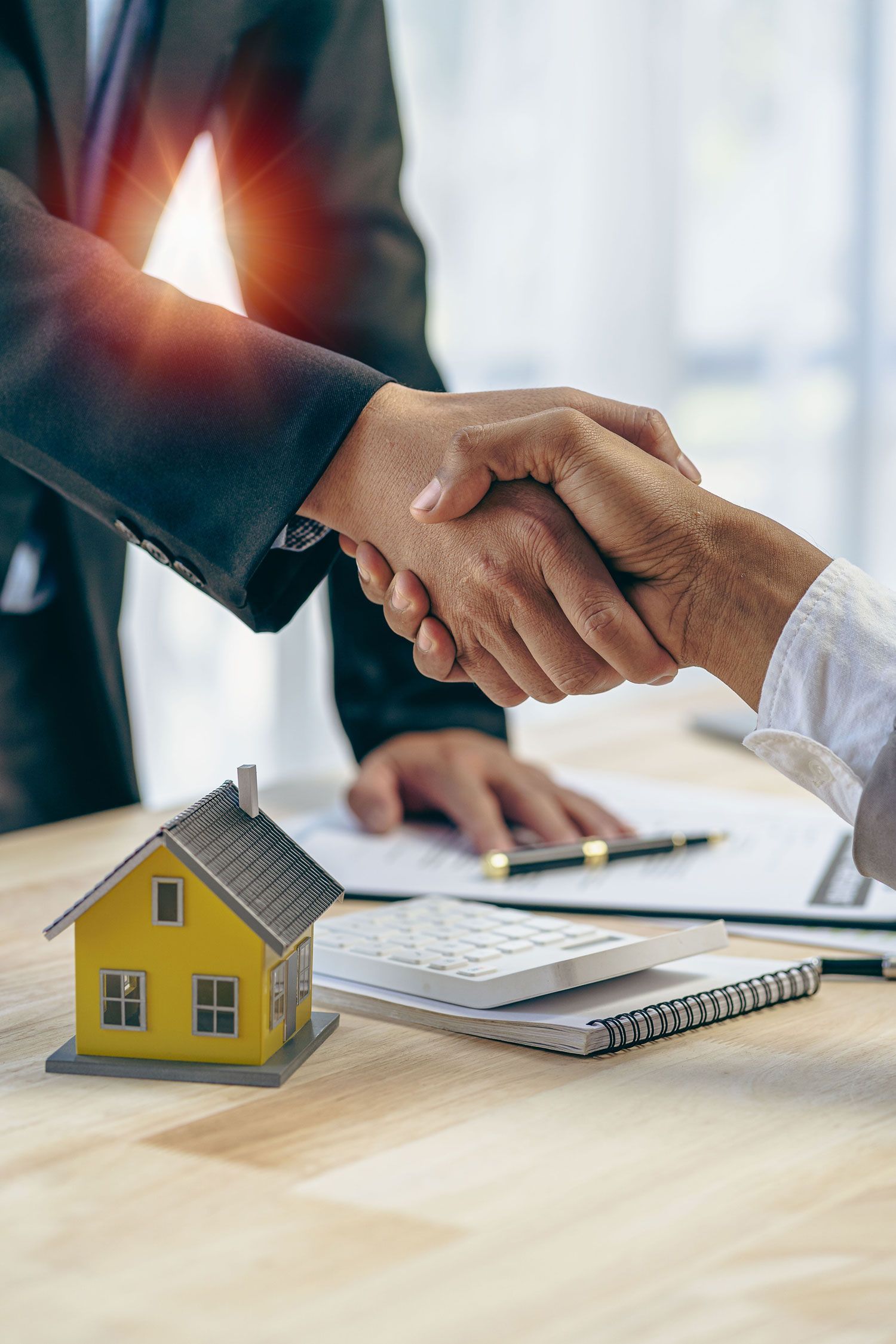 Two people shaking hands over a miniature house and paperwork, symbolizing a real estate deal.