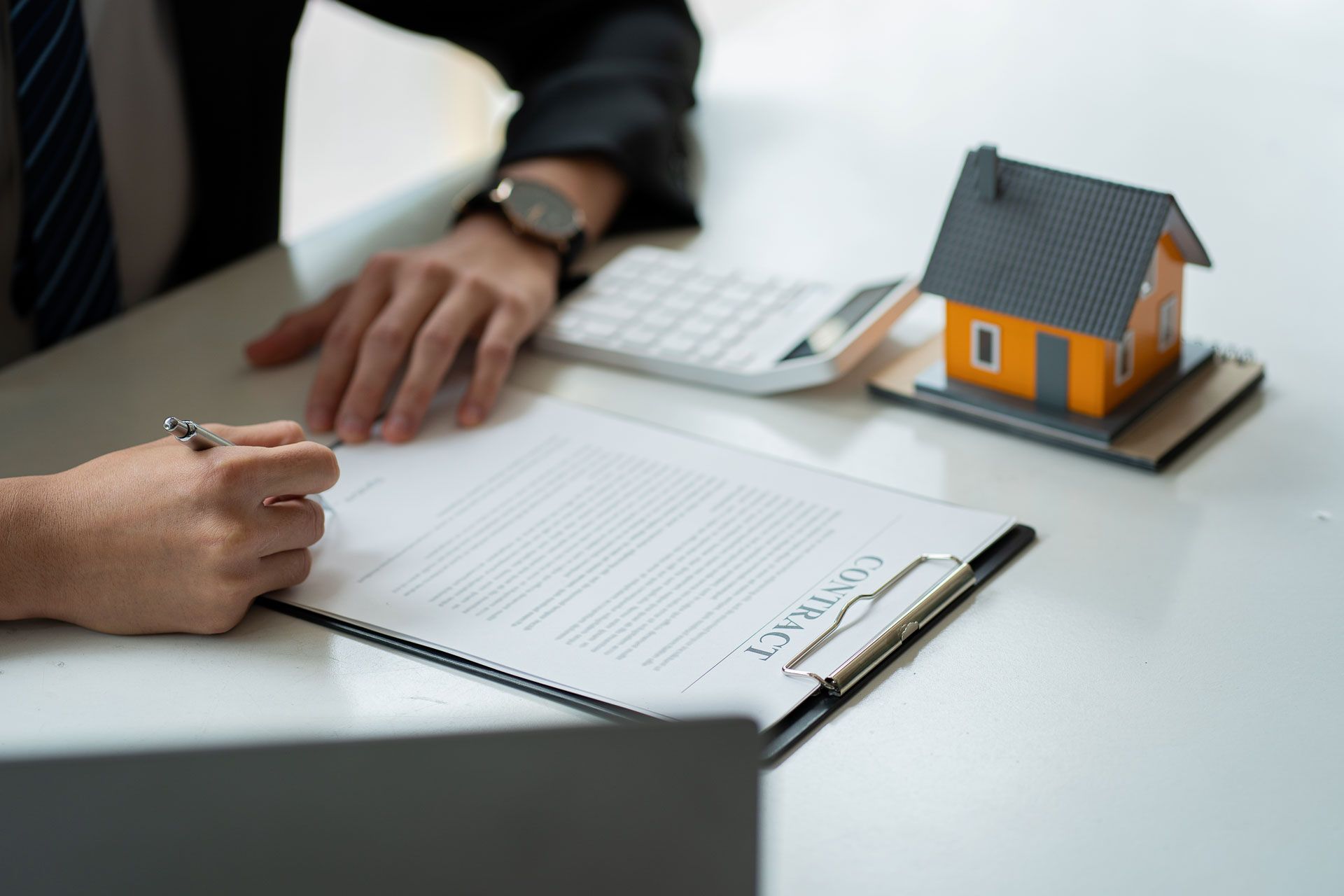 Person in suit signing a contract with a small house model, calculator on the table.