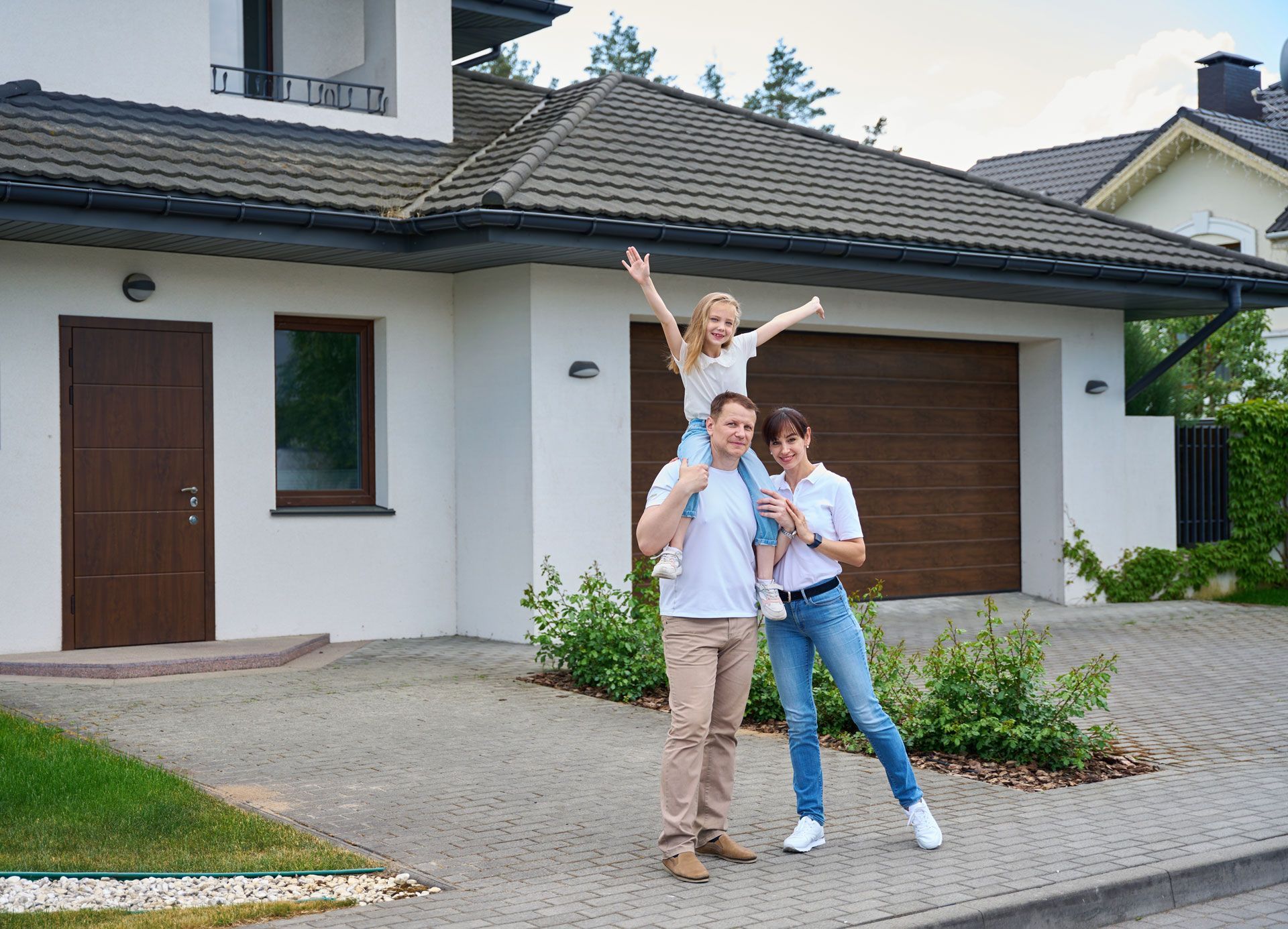 Family of three outside a house; daughter on father's shoulders, arms raised; smiling, paved driveway.