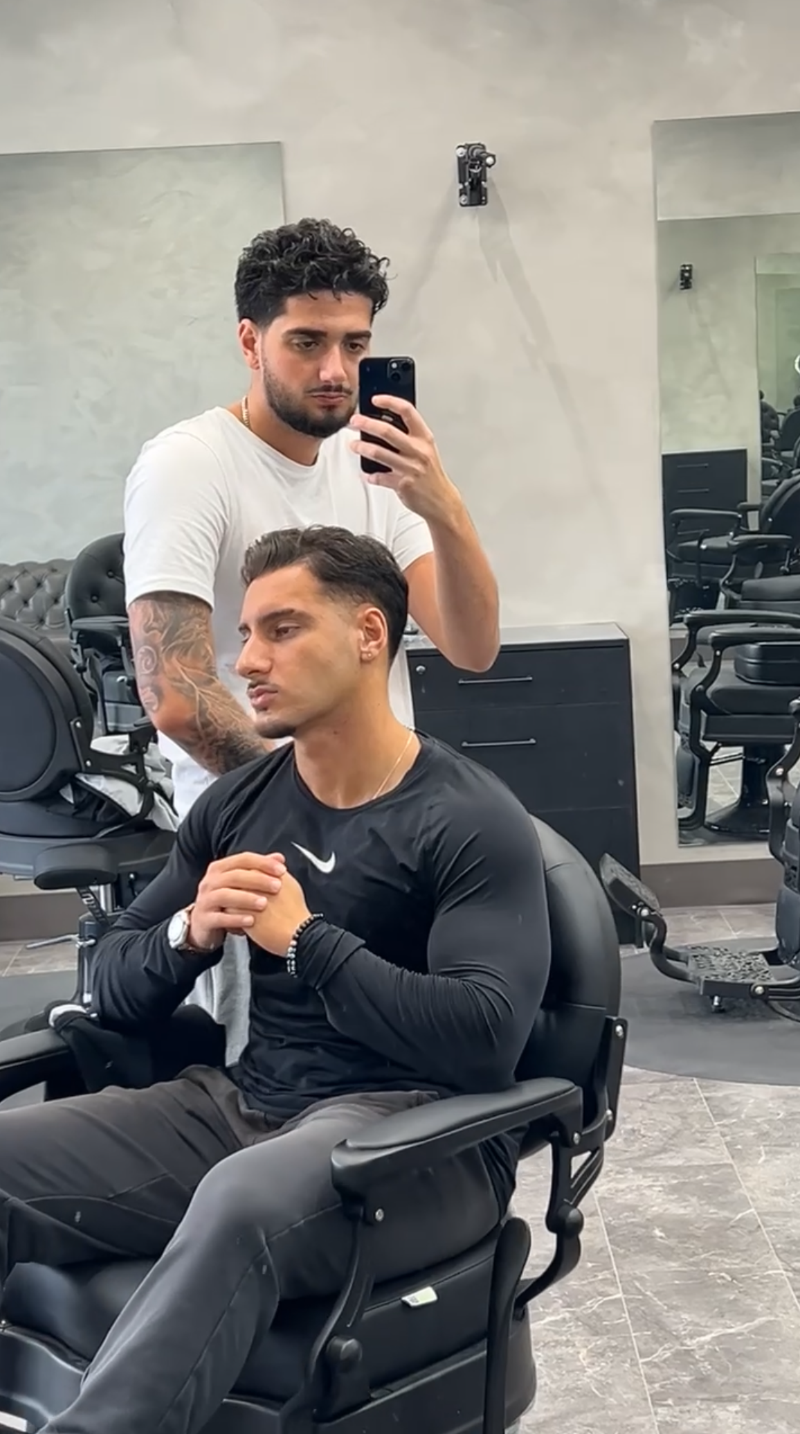 Man getting a haircut at a barbershop. Another man holds a phone up for a selfie in a mirror.