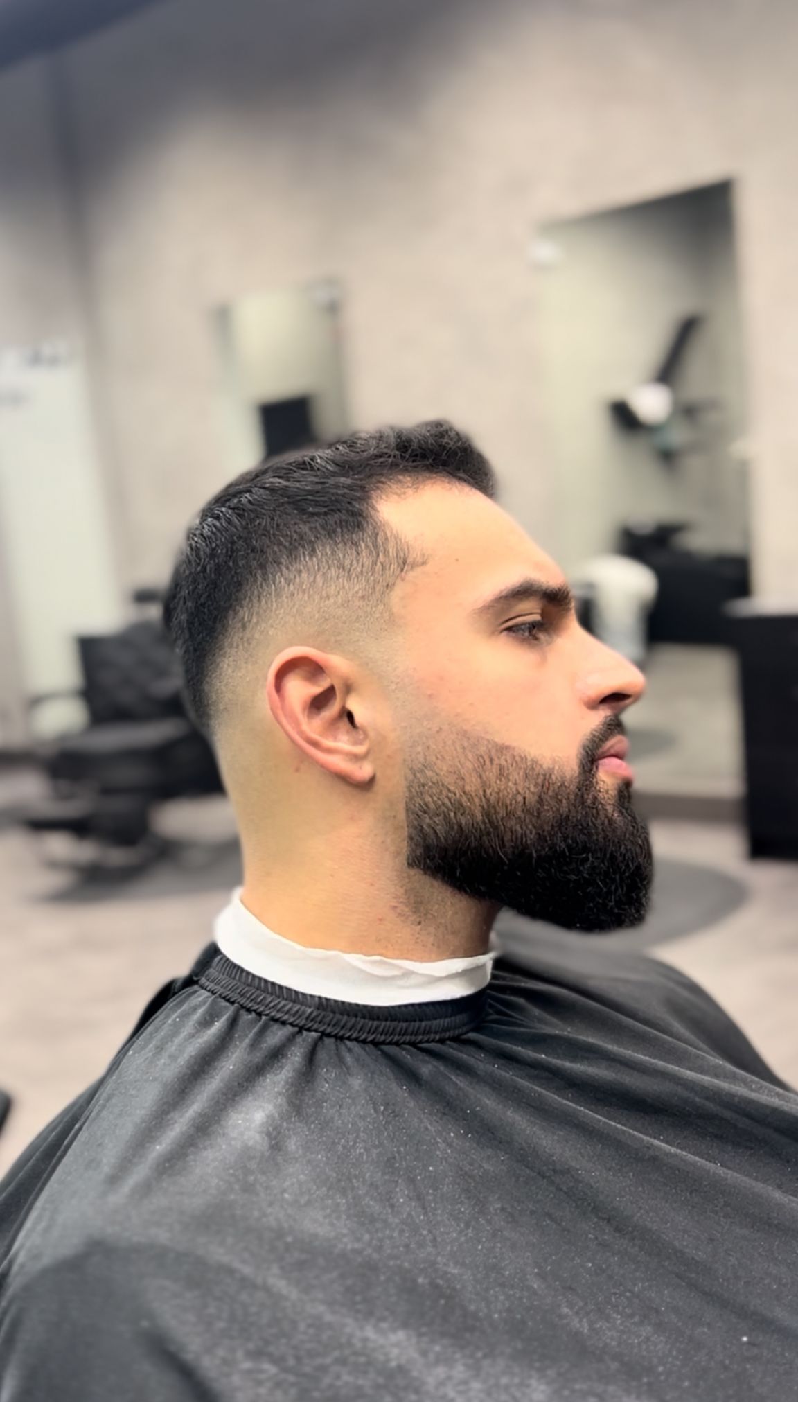 Man with faded haircut and beard in a barber shop, wearing a black cape.