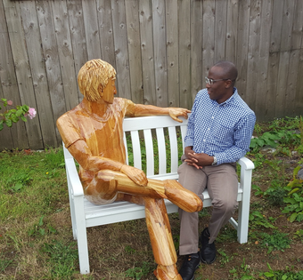 A wooden sculpture of a man sitting next to human man by ingrained Culture