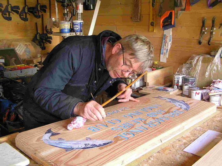 An image of Robert Lawrence, a sculptor and artisan sign maker, painting a wooden sign.