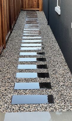 Stone path of rectangular pavers through gravel, beside a wooden fence and dark wall.