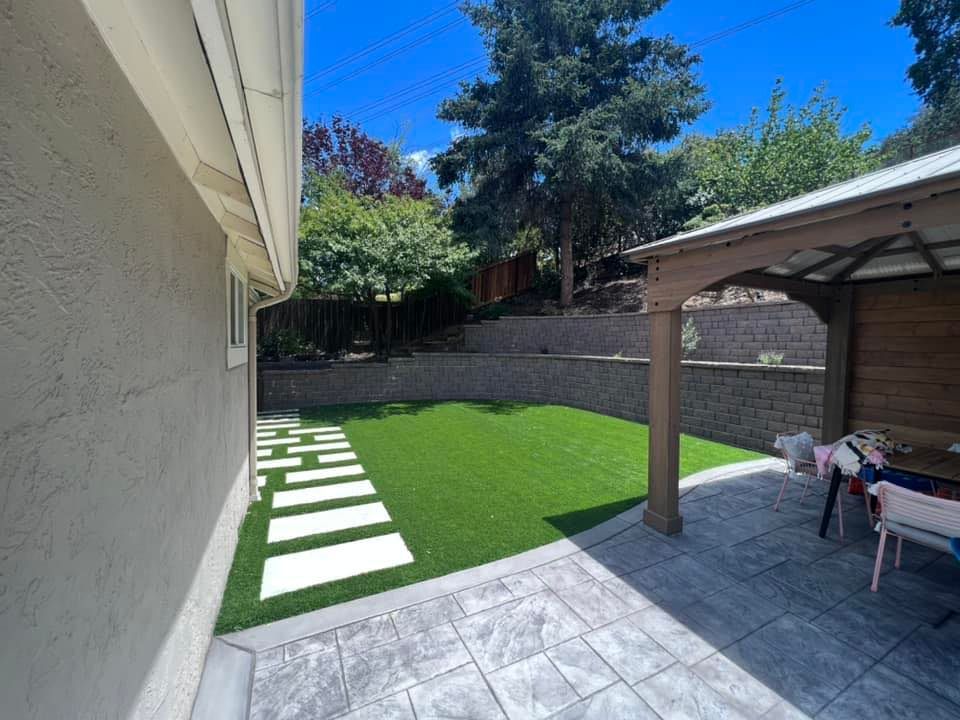 Backyard with artificial turf, stone path, patio, gazebo, and tiered retaining wall. Sunny day.