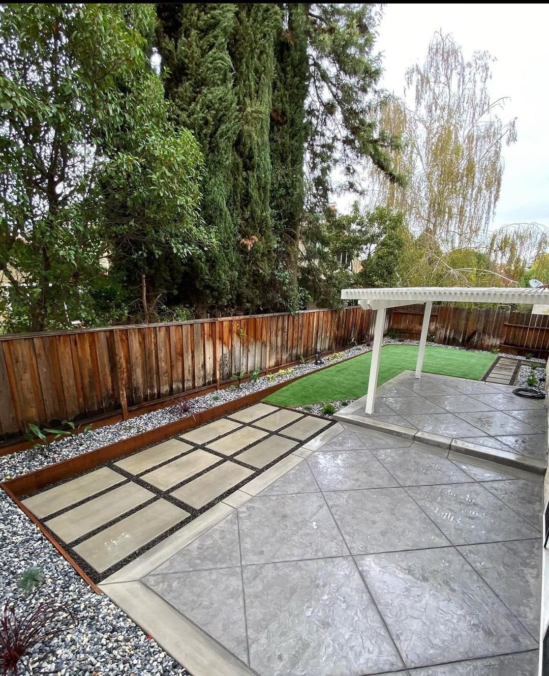 Backyard with concrete patio, lawn, wooden fence, and various trees.