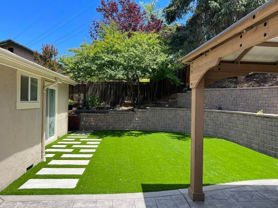 Backyard with green turf, stone pathway, wooden pergola, and retaining wall.