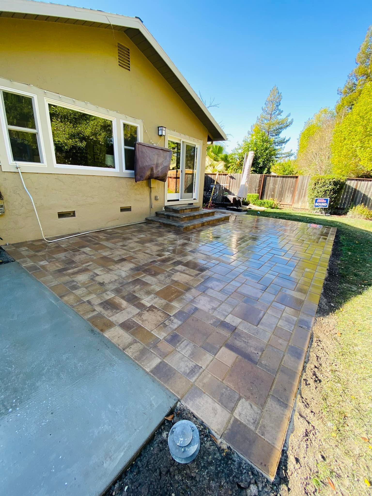 Backyard patio with brick pavers and concrete slab, adjacent to a beige house with a sunny sky.