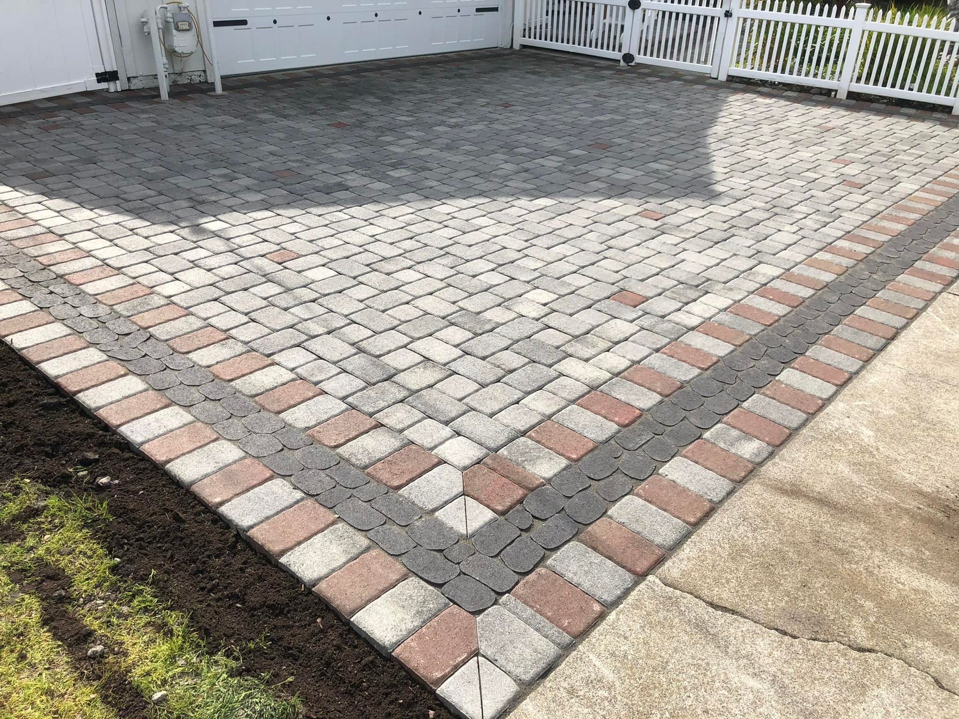 Brick driveway with grey and red border in front of a white garage and fence.