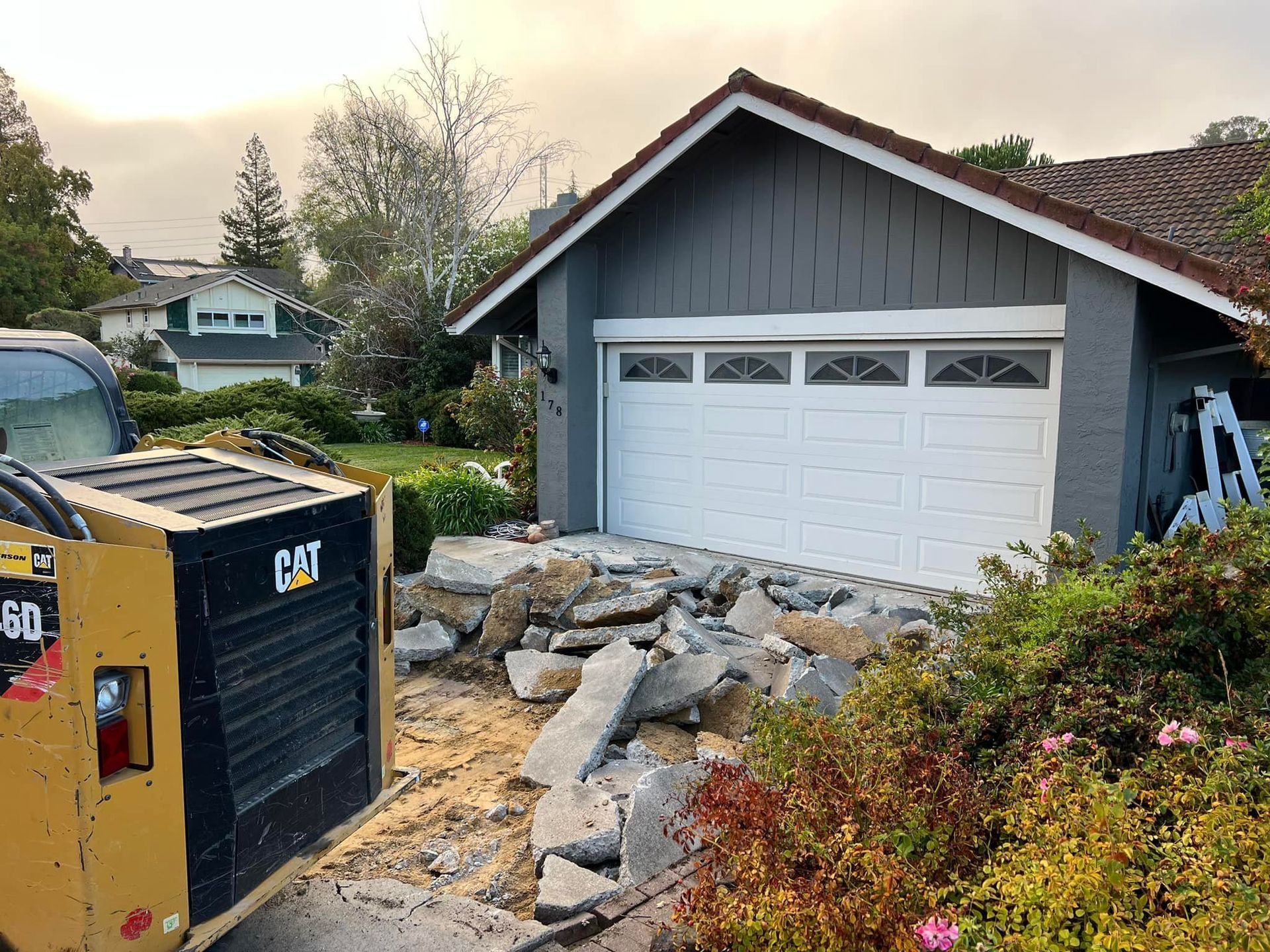 A garage under construction with a CAT excavator in the foreground. Gray garage door, broken concrete, and plants.