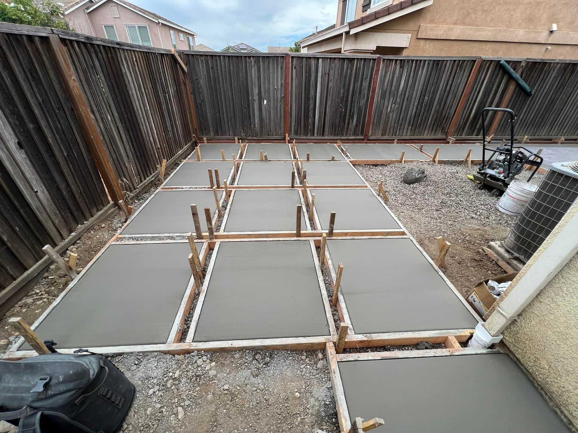 Freshly poured concrete slabs in a backyard, framed by wooden forms, next to a fence and a house.
