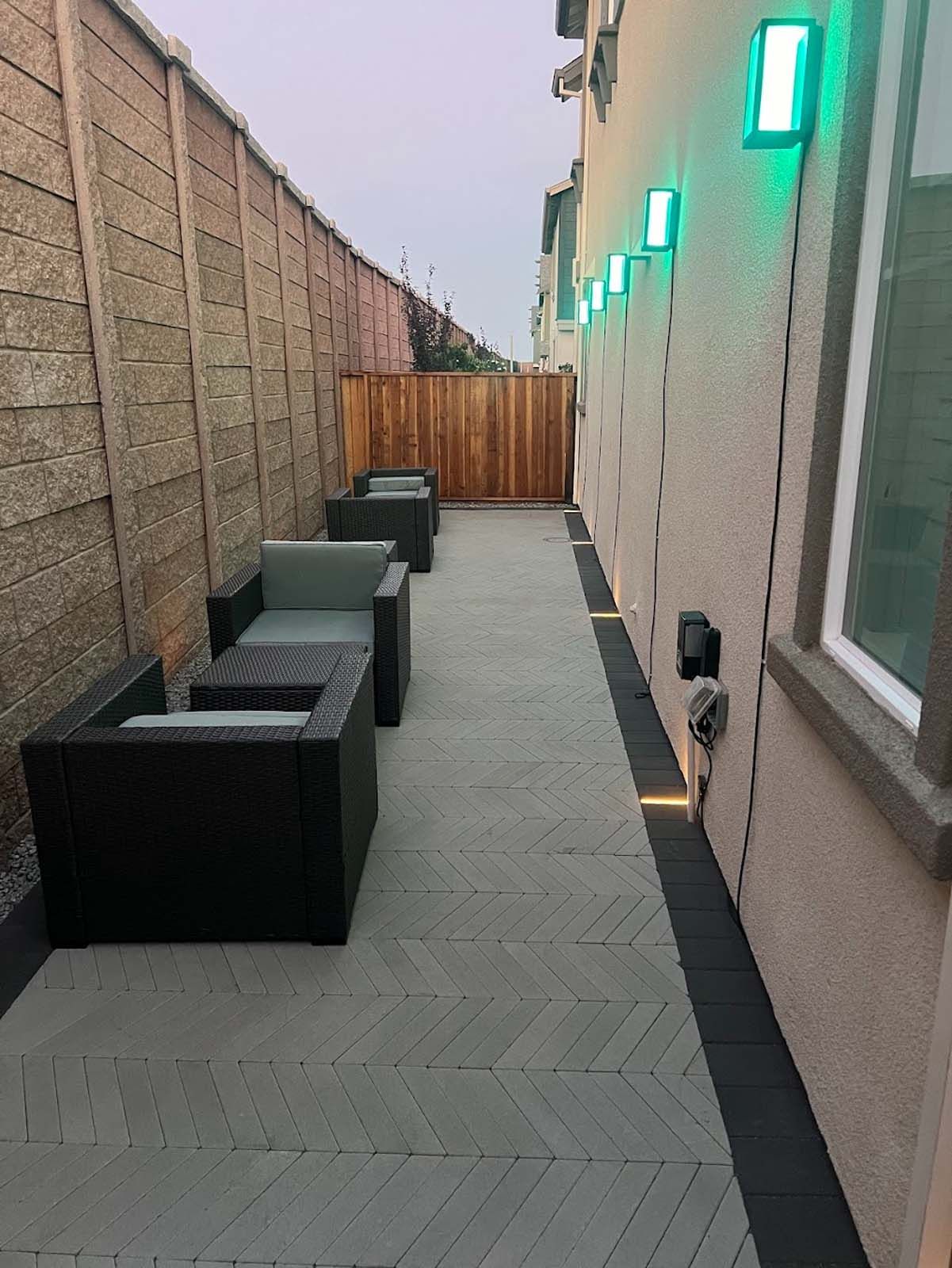 Narrow outdoor patio with modern furniture between a wall and a building, lit by green lights.