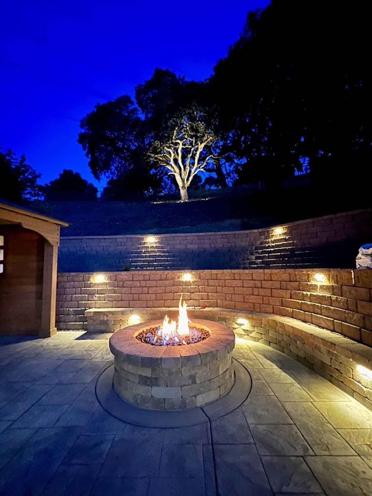 Fire pit on a patio at night with built-in seating, lit by spotlights. A tree is illuminated in the background.