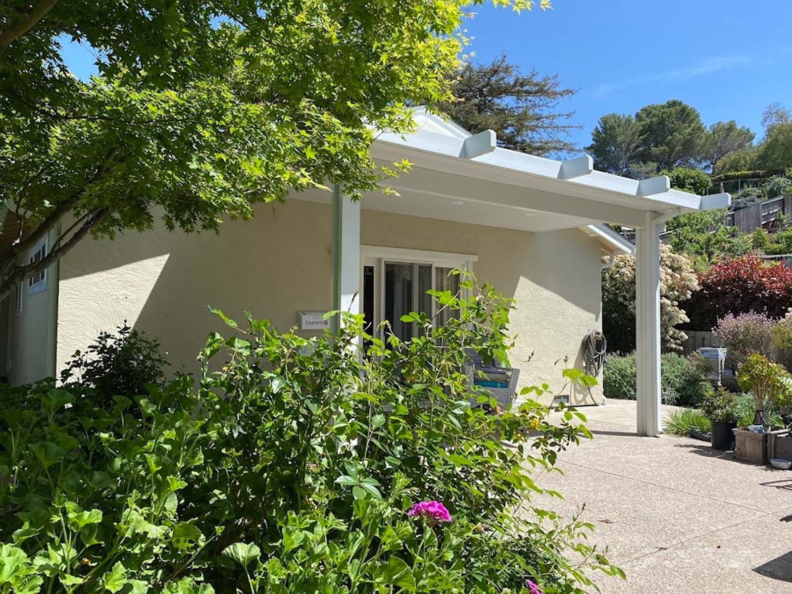 Cream-colored building with pergola, surrounded by lush green plants and a sunny outdoor setting.