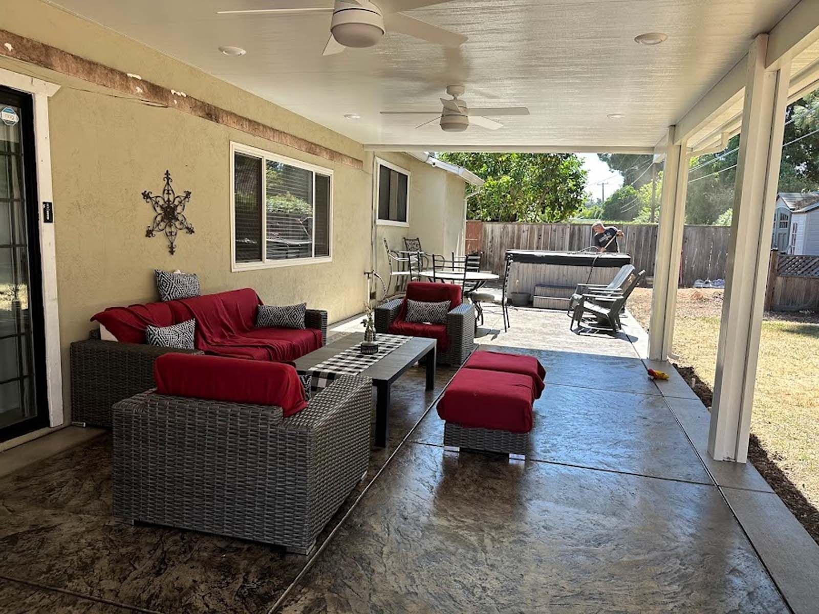 Covered patio with gray wicker furniture, red cushions, and a hot tub in the backyard.