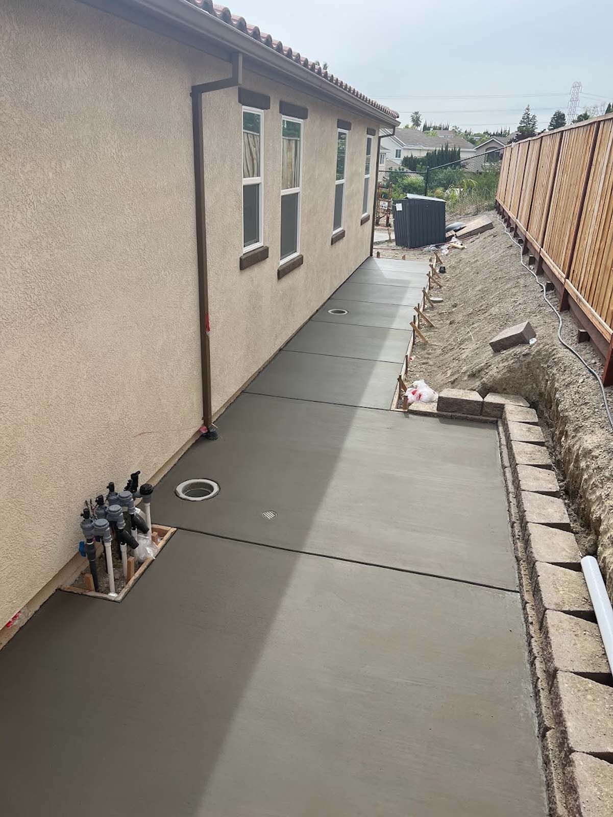 Newly poured concrete walkway along a beige house, with a brick border and a fence.