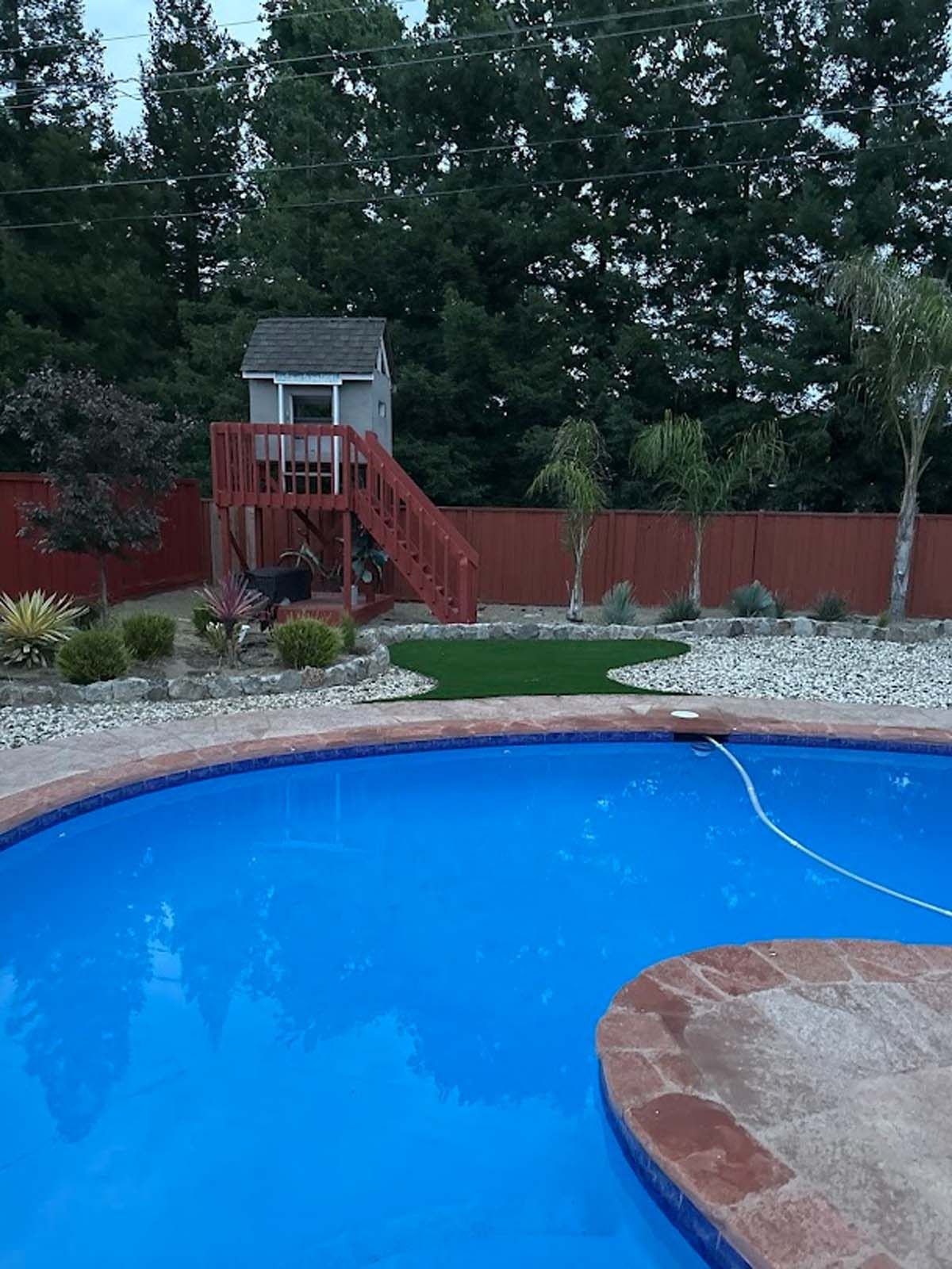 Pool with wooden stairs leading to a playhouse. Red fence, trees, and shrubs in the background.