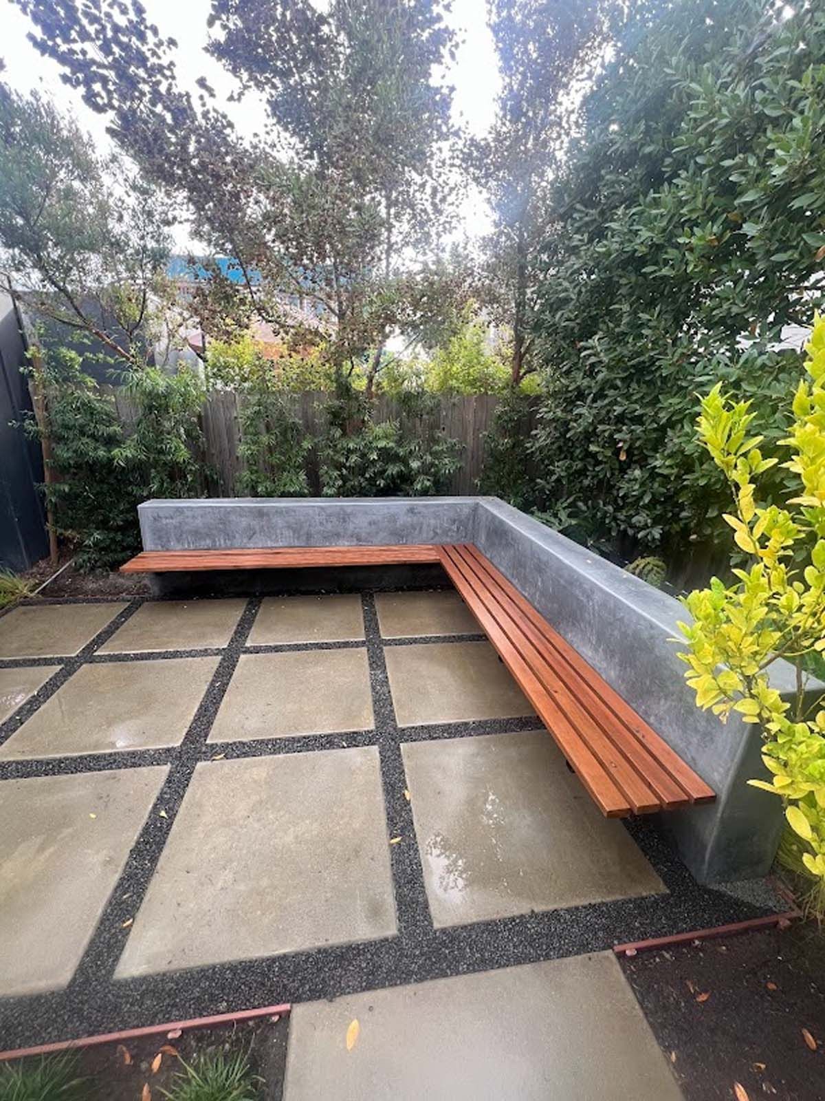 L-shaped outdoor concrete bench with wooden slats on a tiled patio surrounded by greenery.