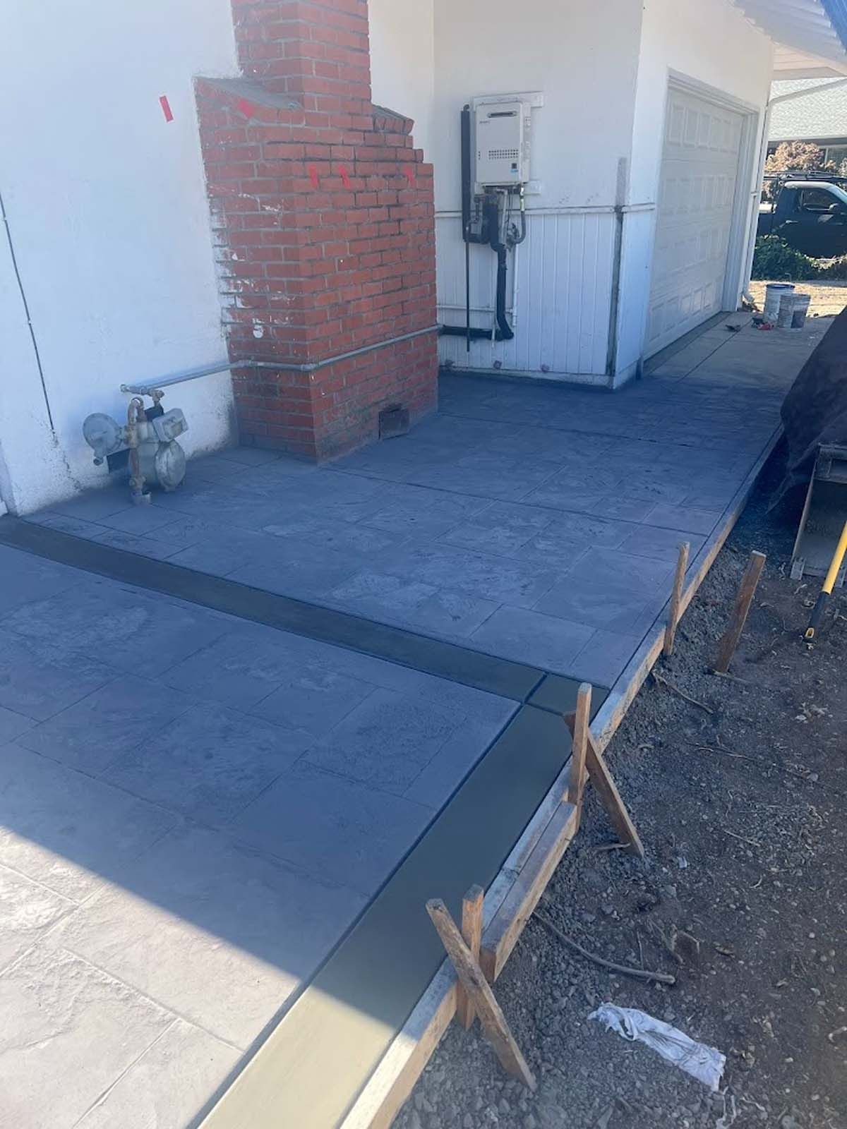 Concrete pathway being poured with wooden forms set up. Brick chimney and white building visible.