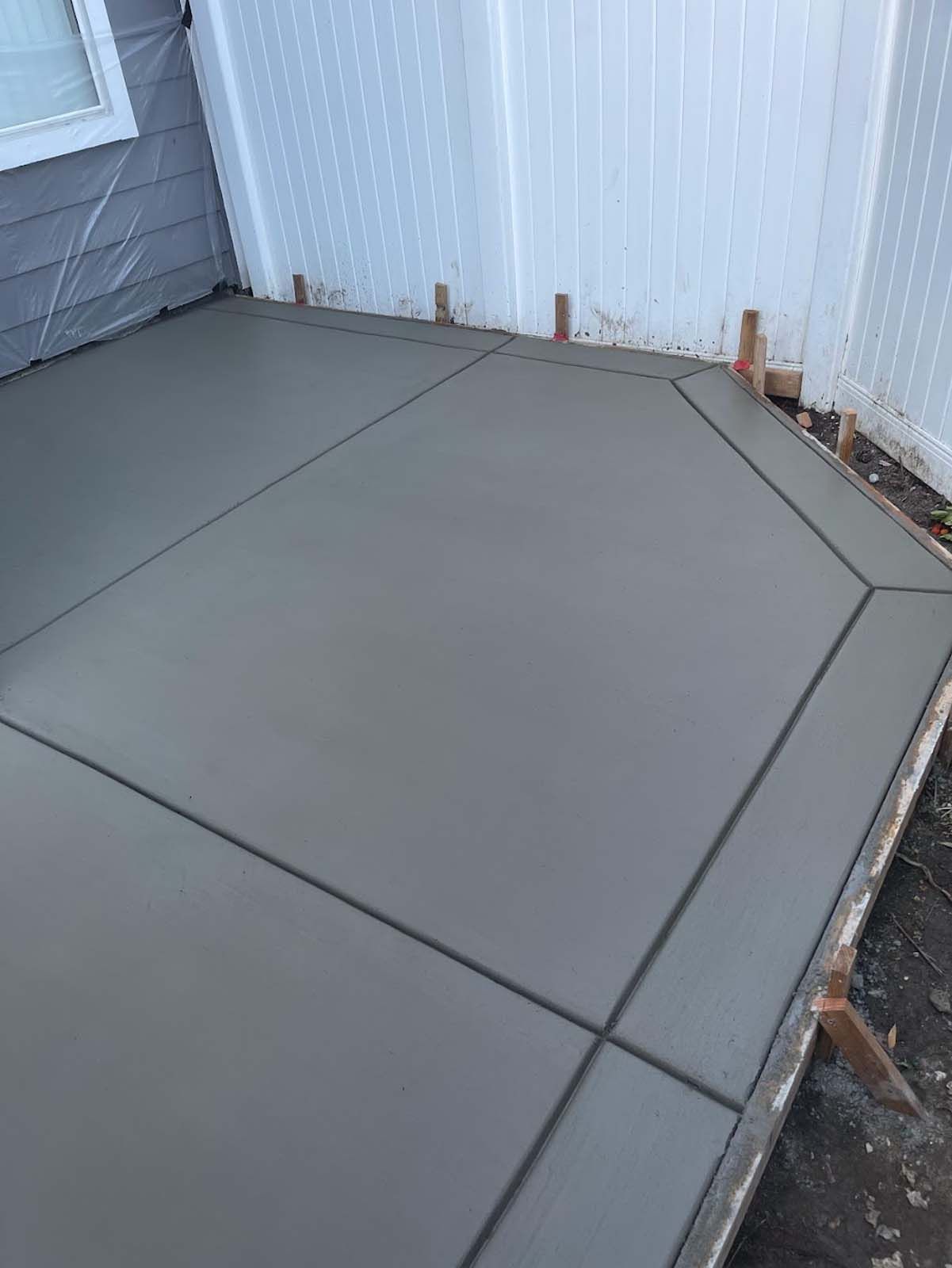 Freshly poured gray concrete patio with geometric design, next to a white fence.