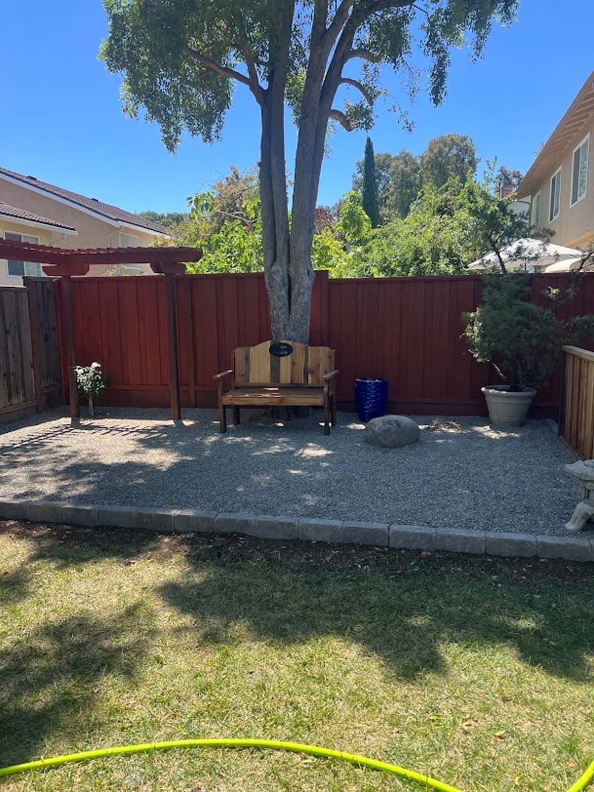 Backyard with gravel area, red fence, wooden bench around a tree, and green grass.