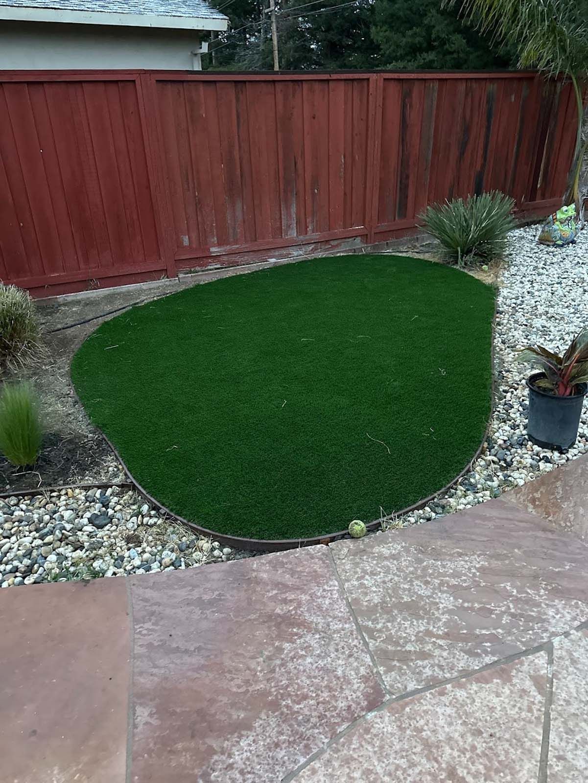 Green artificial turf in a pebble-surrounded bed, next to a patio, with a red wooden fence in the background.