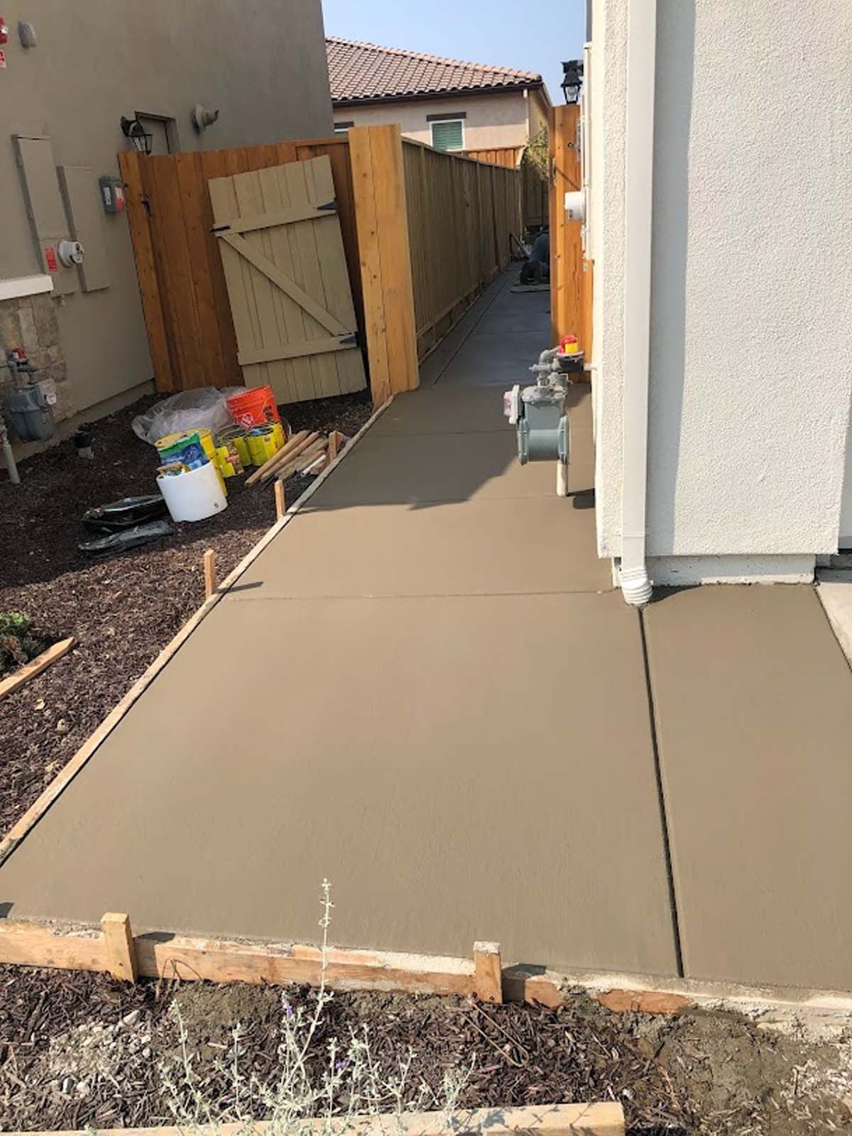 Newly poured concrete walkway next to a house and wooden fence, with wooden borders and construction supplies.