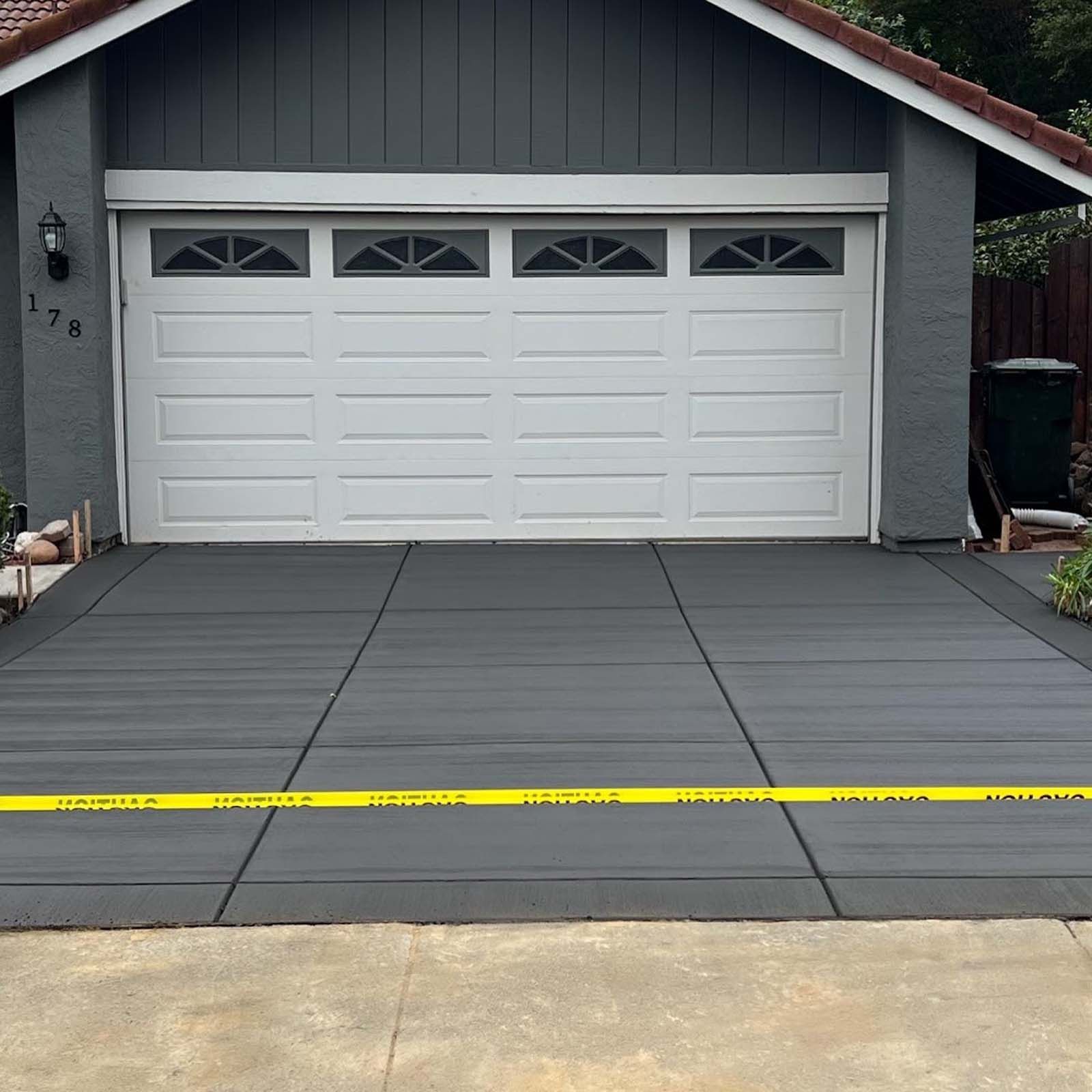 Newly poured dark gray concrete driveway in front of a white garage. Yellow caution tape visible.