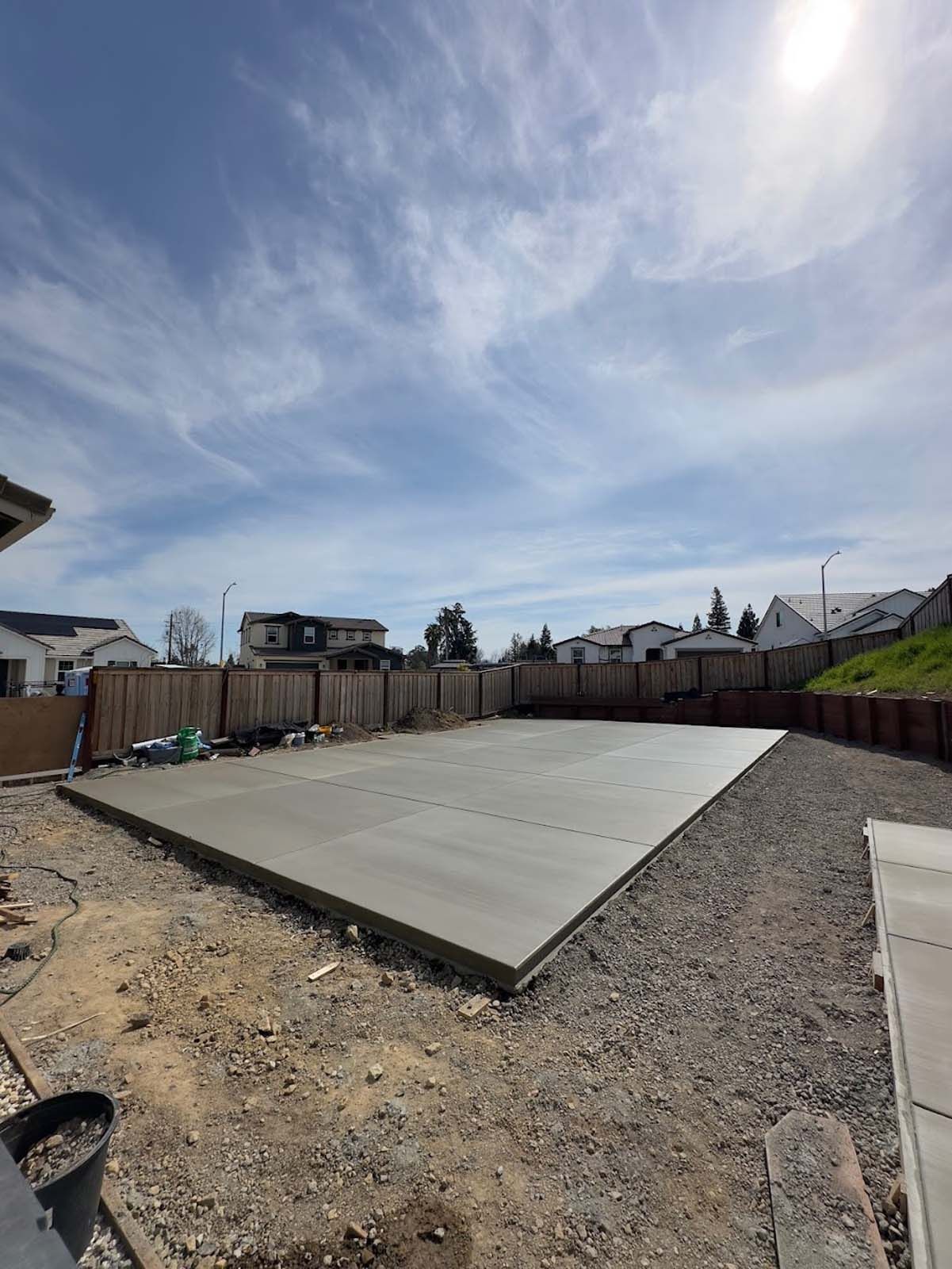 Newly poured concrete slab in a backyard, surrounded by gravel, fence, and houses under a cloudy sky.