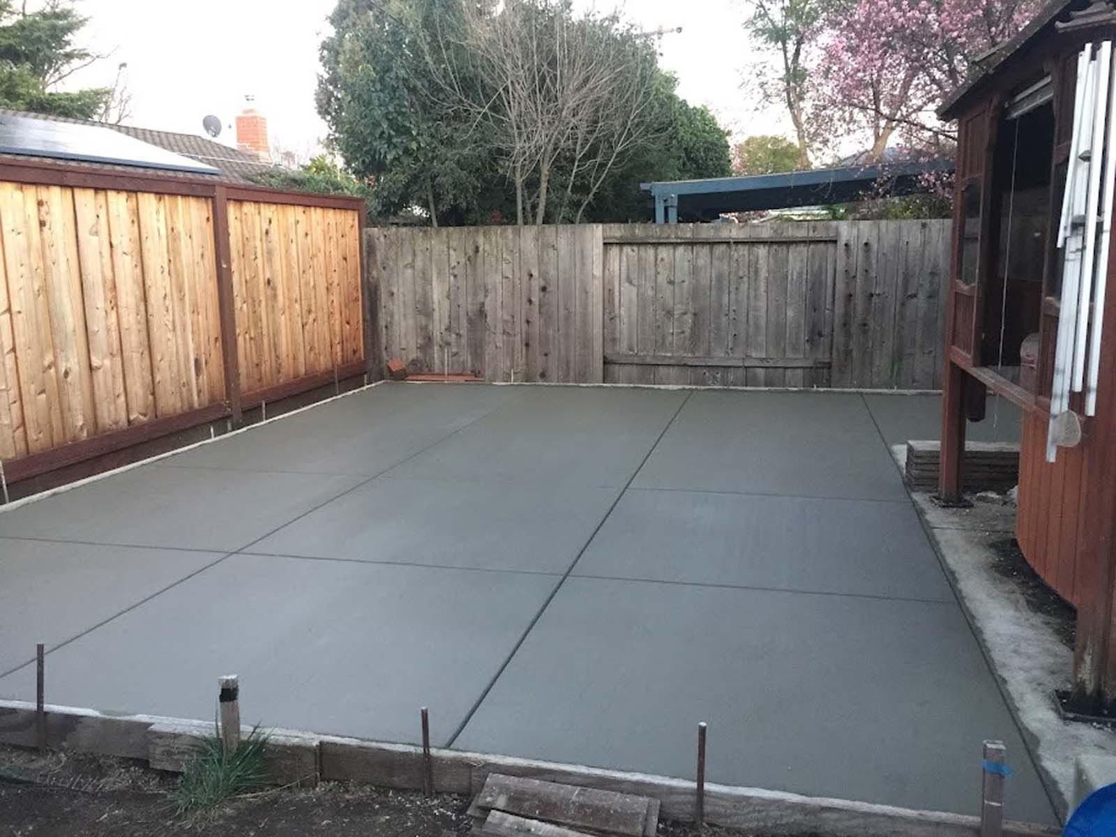 Freshly poured gray concrete patio in a backyard, bordered by wooden fences and a small wooden structure.