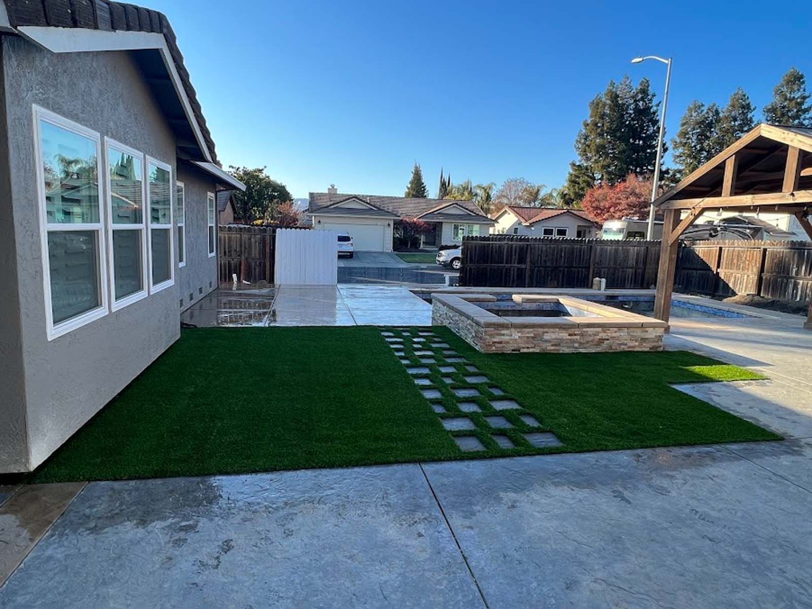 Backyard with green turf, concrete pavers, fire pit, and gazebo under a blue sky.