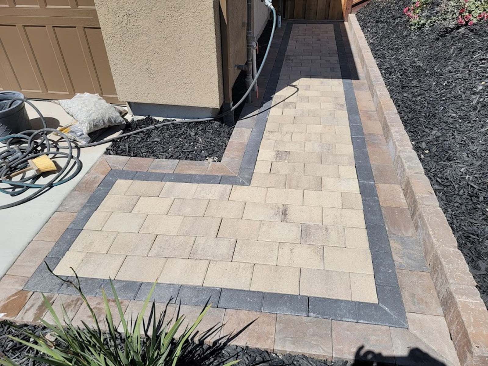 Stone walkway with border, leading to a doorway, adjacent to a garden with dark mulch.