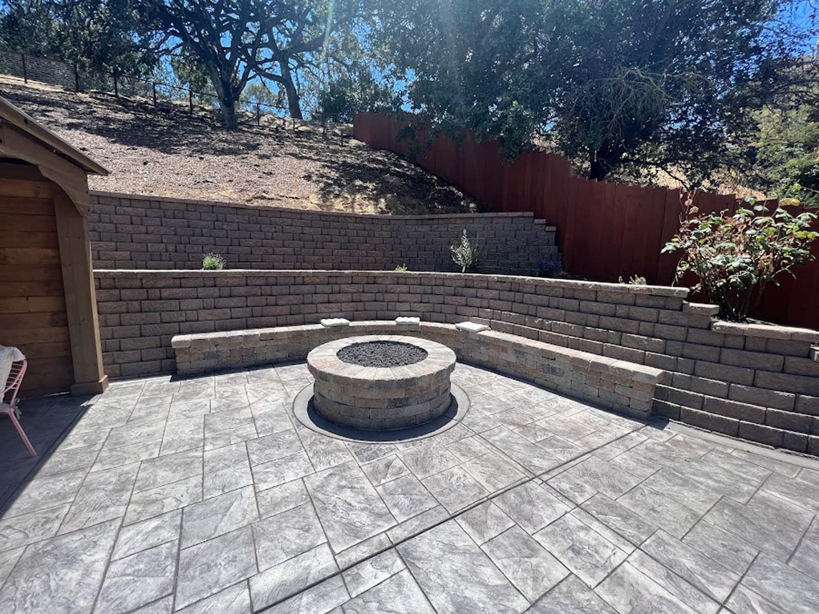 Patio with fire pit, retaining walls, and benches, on a hillside. Gray paving stones, blue sky.