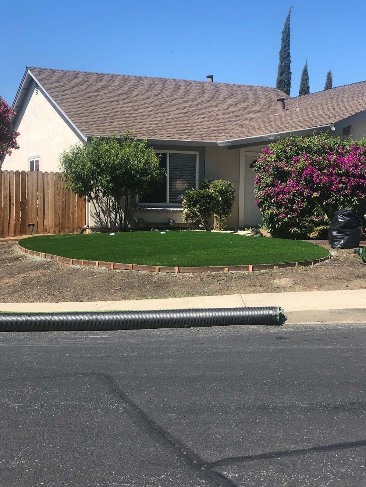 House with green lawn, brown roof and trim, trees, and a wooden fence under a blue sky.
