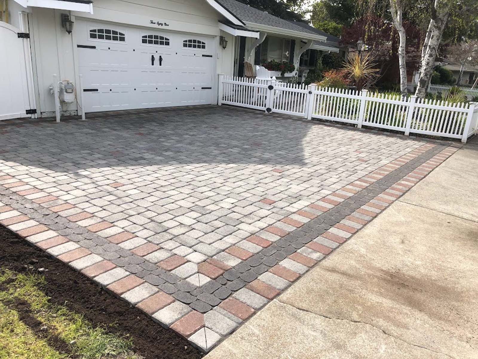 Brick driveway with red, gray, and black pavers, bordering a white picket fence and a garage.