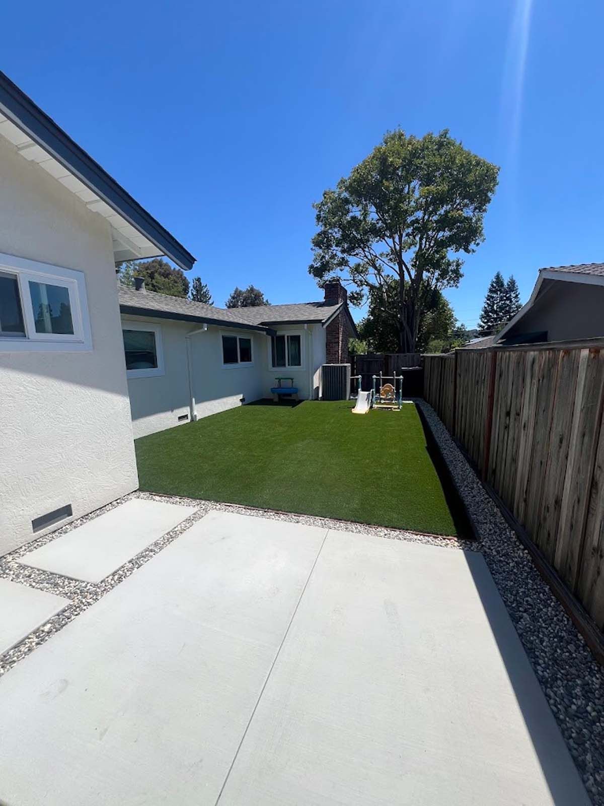 Backyard with green turf, concrete patio, wooden fence, and a house under a sunny blue sky.