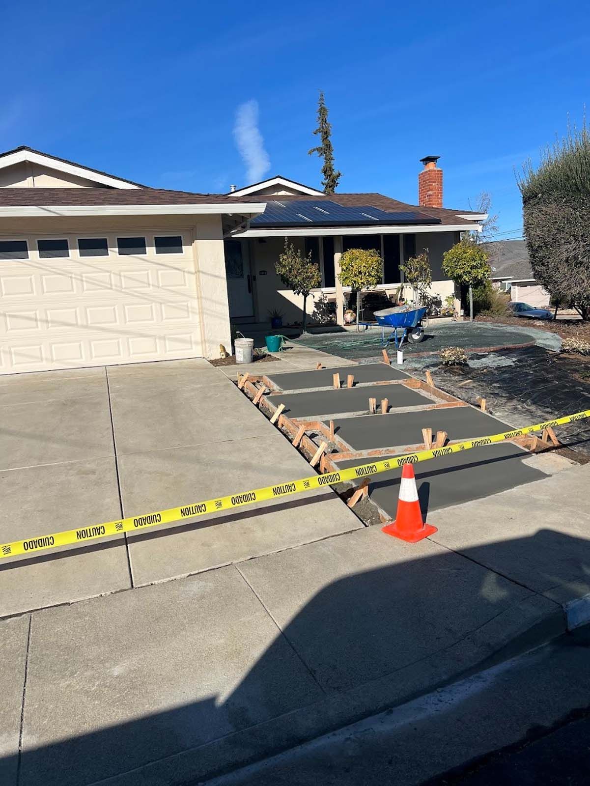 Concrete driveway under construction in front of a beige house with a blue sky. Orange cone, yellow tape.