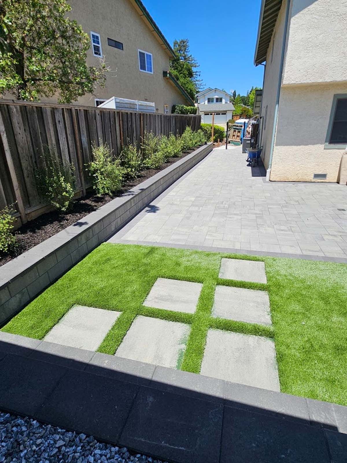 Backyard with a turf area, stepping stones, and a paved walkway next to a fence and house.