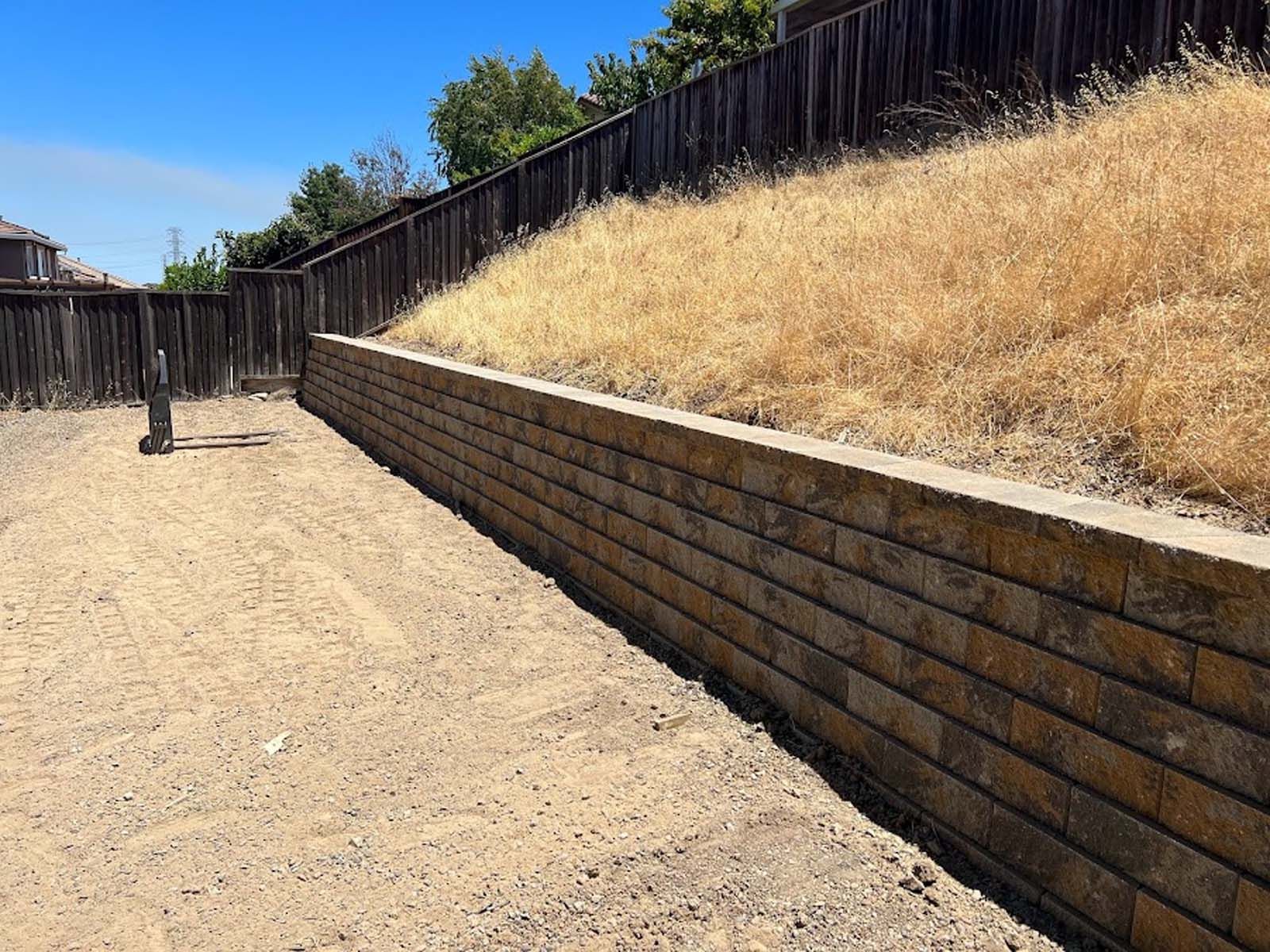 A long retaining wall built of tan bricks separates a gravel yard from a dry, grassy hillside.