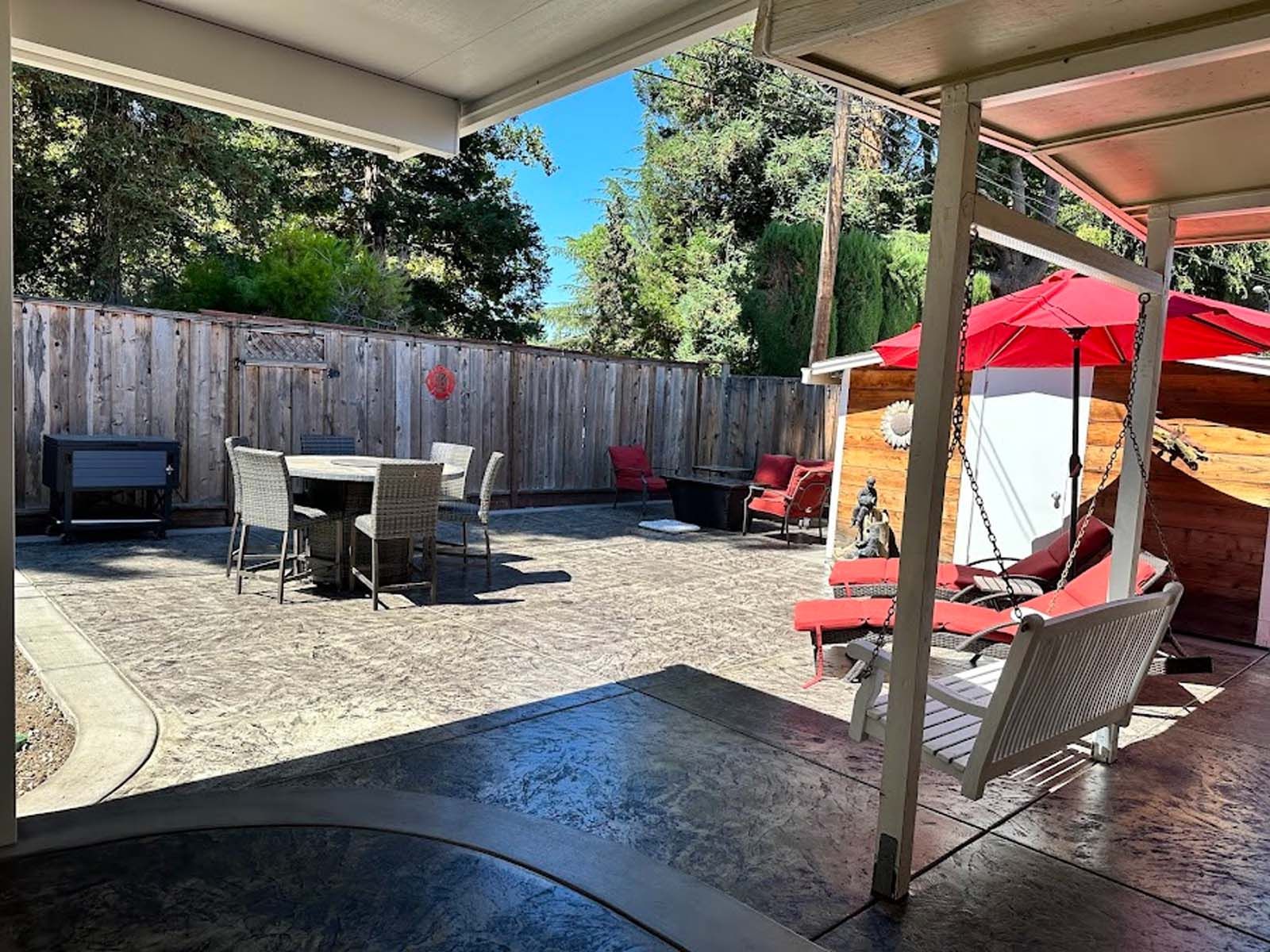 Patio with table, chairs, swing, red umbrella, and wooden fence in the background under a sunny sky.
