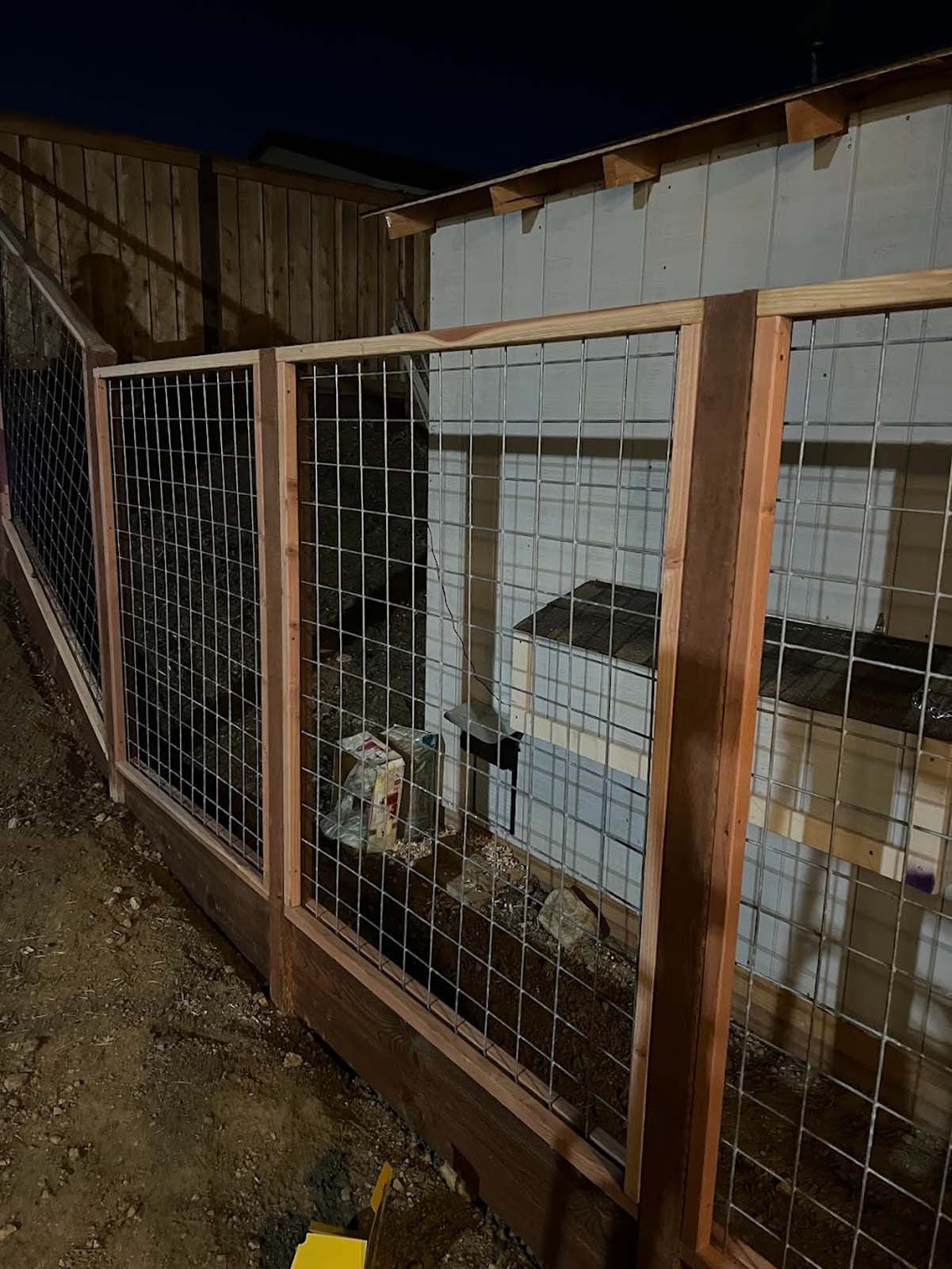 Wooden fence with wire mesh panels surrounding a small, white building at dusk.