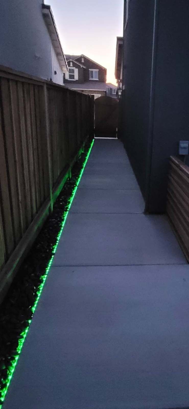 A narrow pathway with green lights along a wooden fence. Buildings are on either side, with the sky visible ahead.