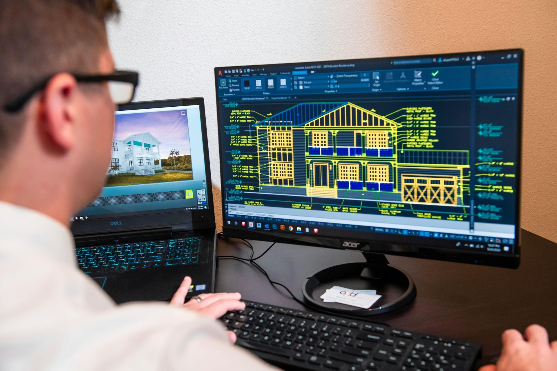 Man working on architectural designs at a desk with a computer and a laptop, showing a house blueprint.