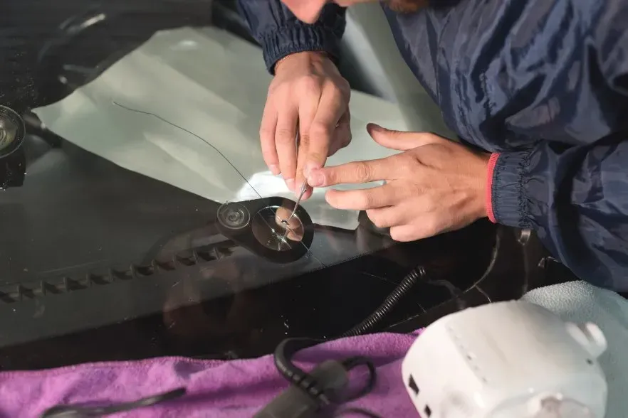 Person in blue coveralls using tools to work on vehicle's windshield near a sensor.